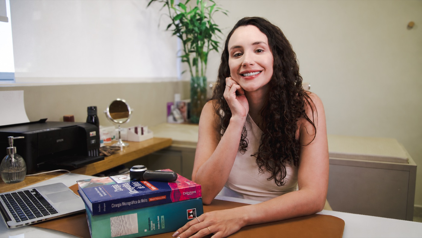 Dra. Carolina Inka em seu consultório, com os livros de Cirurgia Micrográfica de Mohs e o dermatoscópio sobre a mesa
